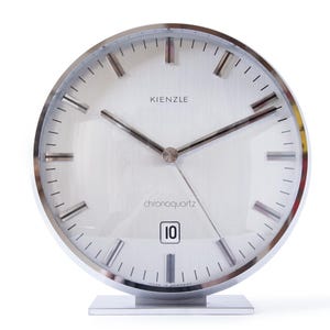 May include: A silver-toned analog clock with a white face and black hands. The clock has a chrome frame and a silver base. The clock face has the text "Kienzle chronoquartz" and the number "10" displayed.