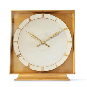 May include: A gold-toned square clock with Roman numeral markings on a white face. The clock has a gold-toned base and hands.