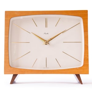 May include: A vintage wooden clock with a white face and gold hands. The clock is made by the company "Nautica" and says "Made in Germany" on the face. The clock has a square shape with rounded corners and sits on four small legs.