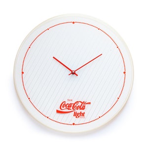 Puede incluir: Reloj redondo blanco con agujas rojas y un logotipo rojo de Coca-Cola Light. La esfera del reloj tiene un fondo blanco con puntos rojos alrededor del borde y finas líneas rojas.