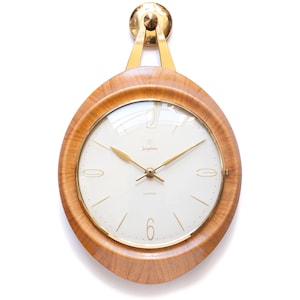 May include: A vintage wall clock with a light wood frame and a white face. The clock has gold-colored hands and a gold-colored mounting bracket. The clock face has the brand name "Junghans" printed on it.