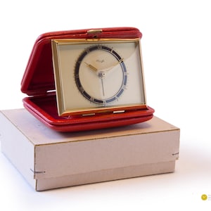 Può includere: Una custodia da viaggio in pelle rossa con un orologio da sveglia dorato all'interno. Il quadrante dell'orologio ha indici orari in numeri romani neri e lancette nere.