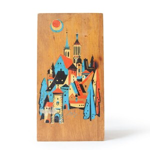 May include: Wooden plaque featuring a colorful, stylized illustration of a European town with towers, buildings, and trees. The artwork includes a sun and is painted in shades of orange, blue, and black.