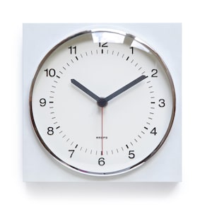 Op de afbeelding: Een witte vierkante klok met een ronde wijzerplaat. De wijzerplaat is wit met zwarte cijfers en wijzers. De klok heeft een zilverkleurige rand en de merknaam "KRUPS" staat onderaan. De secondewijzer is rood.