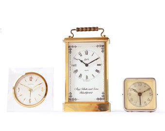May include: Three vintage clocks, one with a clear acrylic case, one with a brass case, and one with a silver case. The brass clock has a white face with Roman numerals and the text "Quartz" and "Aug. Schatte and Sons Blackforest".