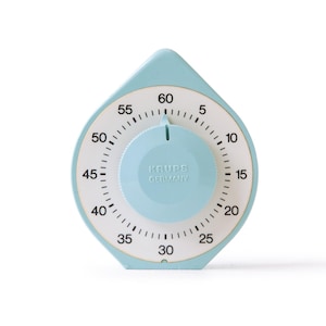 Puede incluir: Un temporizador de cocina de color azul claro en forma de gota con una esfera circular blanca que muestra números negros del 5 al 60. El dial central es azul claro y presenta el texto en relieve KRUPS GERMANY. Tiene una parte superior puntiaguda y base plana.