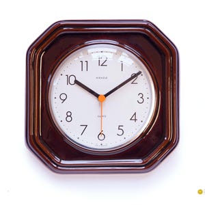 Könnte beinhalten: Eine Vintage Kienzle Quarz-Wanduhr mit einem dunkelbraunen achteckigen Rahmen. Das Zifferblatt ist weiß mit schwarzen Ziffern und Zeigern sowie einem orangefarbenen Sekundenzeiger. Klassisches Design.