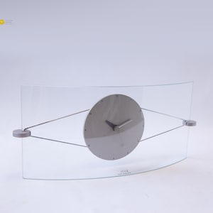 May include: A modern wall clock with a curved glass face and a circular gray clock face. The clock hands and the frame are silver. The clock is suspended by thin metal rods. The text "SESSE DESIGN TOMASZ KRAUS" is printed at the bottom.