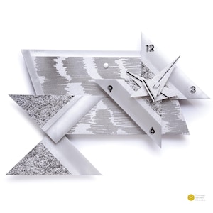 May include: Abstract geometric wall clock with a modern design. The clock features a combination of gray and white triangular shapes, with the numbers 3, 6, 9, and 12 visible. The clock hands are black and white. The clock is a unique piece of home decor.
