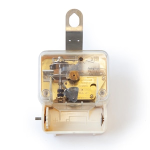 Op de afbeelding: Een vintage uurwerk met een doorzichtige plastic behuizing, metalen componenten en een batterijcompartiment. Het uurwerk is gemarkeerd met "Junghans Germany". De metalen beugel aan de bovenkant maakt ophangen mogelijk. De klok is ontworpen om 24 uur te lopen.