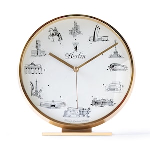 May include: A gold-toned desk clock with a white face featuring black illustrations of Berlin landmarks. The clock face has gold hands and the word "Berlin" in script. The clock is on a gold base.