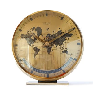 Puede incluir: Un reloj mundial de cuarzo Kienzle vintage con acabado dorado. La esfera del reloj presenta un mapa del mundo y un indicador de zona horaria. El reloj tiene una base rectangular y manecillas doradas.