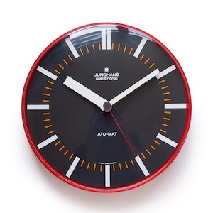 May include: A round, vintage Junghans electronic wall clock with a black face and a red frame. The clock features white hands and markers, with orange accents. The text "JUNHANS electronic" and "ATO-MAT" are visible on the clock face. Made in Germany.