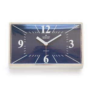 Könnte beinhalten: Rechteckige elektronische Junghans-Uhr mit marineblauem Zifferblatt, weißen Ziffern und weißen Zeigern. Die Uhr hat einen silberfarbenen Rahmen und der Text "JUNGHANS electronic" und "ATO MAT" ist auf dem Zifferblatt zu sehen.