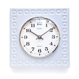 May include: A white ceramic square clock with a silver face and black hands. The clock face has a raised border and the words "EUROPA" and "Clemco" printed on it.
