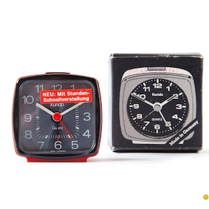 May include: A black alarm clock with a red plastic case. The clock face has white numbers and hands, and the words "Kundo" and "Quartz" are printed on the face. The clock is packaged in a black box with the words "Made in Germany" printed on it.