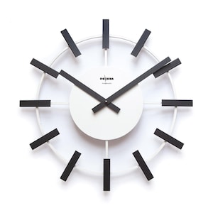 May include: A modern wall clock with a white face and a white circular frame. The clock features black hands and black rectangular hour markers. The text "DEETA HAMBURG" is printed on the clock face. The clock has a minimalist design.