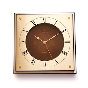May include: Square gold-toned wall clock with a brown face and black Roman numerals. The clock has gold hands and the brand name "Junghans" is visible. The clock is surrounded by a black frame and has gold-colored accents in each corner.