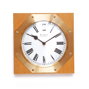 May include: A square wall clock with a light brown wooden frame and an octagonal brass face plate secured by eight screws. The white clock face features black Roman numerals, black hands, and the text WEIMAR QUARTZ printed in black.