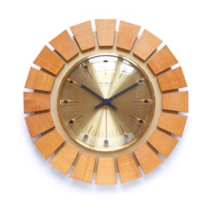 May include: A round, mid-century modern wall clock with a gold-tone face and black hands. The clock is surrounded by a ring of light brown wooden blocks. The clock face has the word "WEIMER" printed on it.