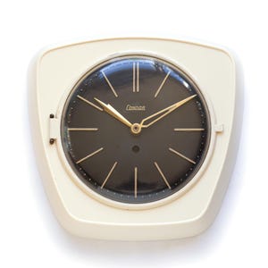 Könnte beinhalten: Eine Wanduhr mit einem cremefarbenen, trapezförmigen Kunststoffrahmen und einem runden schwarzen Zifferblatt. Die Uhr hat goldfarbene Zeiger und Stundenmarkierungen, wobei der Markenname Condor in goldener Schrift unter der Zwölf steht.
