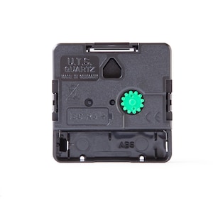 May include: A black plastic U.T.S. Quartz clock movement mechanism with a green adjustment gear. The square housing features molded text including U.T.S. QUARTZ, MADE IN GERMANY, IEC R6, and ABS. It has a central hanger hole and battery compartment.