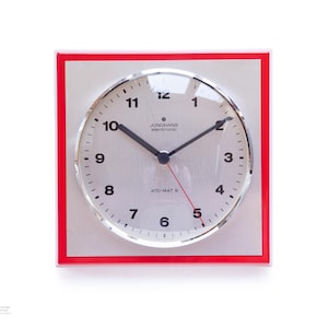 May include: A square, vintage Junghans electronic clock with a silver face and black hands. The clock has a red frame and the text "JUNGHANS electronic ATO-MAT S" is visible. The clock face displays the numbers 1 through 12.