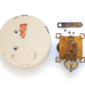 May include: A disassembled vintage clock mechanism with a white clock face. The clock face has a red and white sticker with the text "G.J. GROSS & SON, INC. NEW YORK". The clock mechanism is made of brass and has a winding key and a pendulum.