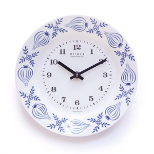 Könnte beinhalten: Weiße Keramik-Wanduhr mit blauem Zwiebel- und Blumendesign am Rand. Das Zifferblatt hat schwarze Zeiger und den Text "WEIMAN". Die Uhr ist mit "MADE IN JAPAN" gekennzeichnet.