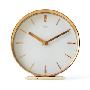 Könnte beinhalten: Eine vintage goldfarbene elektrische Uhr mit weißem Zifferblatt. Die Uhr hat goldene Zeiger und Markierungen, mit dem Markennamen "Kienzle Electric" sichtbar. Die Uhr ist rund und steht auf einem rechteckigen Sockel.