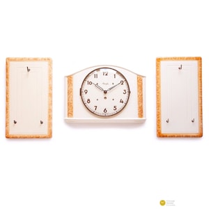 Könnte beinhalten: Eine weiße Vintage-Uhr mit braunem Zifferblatt und schwarzen Zeigern. Die Uhr ist an einer weißen Wand montiert, mit zwei weißen rechteckigen Paneelen mit braunen Verzierungen auf beiden Seiten.