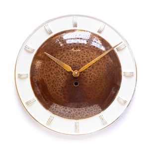 Könnte beinhalten: Eine runde Wanduhr mit einem weißen Außenring und einem strukturierten braunen Zentrum. Die Uhr hat goldfarbene Zeiger und römische Ziffern als Stundenmarkierungen. Das Zifferblatt trägt den Schriftzug "Marvin" in Gold.