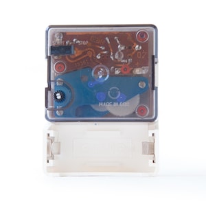 May include: A vintage clock movement with a clear plastic cover, revealing internal gears and circuitry. The movement features a blue and silver oscillating mechanism, red accents, and a white battery compartment. The text "START" and "STOP" are visible.
