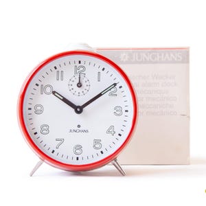 Peut inclure: Un réveil Junghans vintage rouge et blanc avec un cadran blanc, des aiguilles noires et un petit cadran des secondes. L'horloge a un bord rouge et des pieds argentés. La boîte d'origine est visible en arrière-plan.