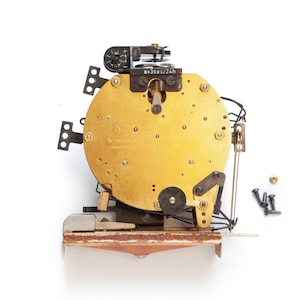 May include: A vintage clock movement with a gold-colored circular face and various mechanical components. The clock mechanism includes a small dial with the text "+3sec/24h" and several screws.
