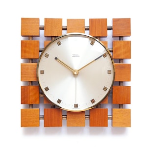 Könnte beinhalten: Eine quadratische Wanduhr mit hellem Zifferblatt und goldfarbenen Zeigern und Zierleisten. Die Uhr ist von einem Gitter aus Holzblöcken und Metallstäben umgeben. Auf dem Zifferblatt steht "Diva Electric".