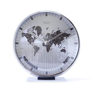 May include: A round Kienzle Quartz world map clock with a silver metallic finish. The face features a black world map, thin silver hands, and a curved 24-hour time scale at the bottom. The clock is mounted on a rectangular silver base stand.