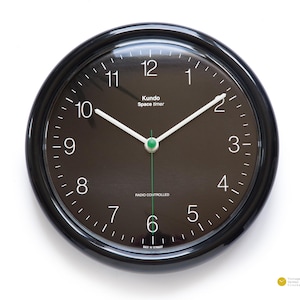 Op de afbeelding: Een zwarte ronde klok met witte cijfers en wijzers. De wijzerplaat van de klok is zwart met een groene secondewijzer. De wijzerplaat van de klok heeft ook de tekst "Kundo Space timer" en "RADIO CONTROLLED".
