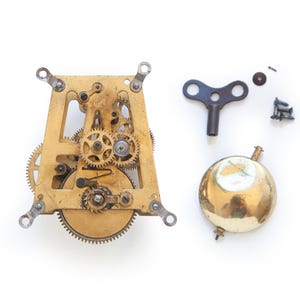 May include: A disassembled clock mechanism with a brass frame, gears, and a bell. A dark metal key, screws, and a small washer are also present. The clock parts are arranged on a white surface.