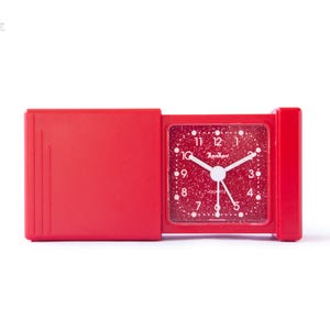 Op de afbeelding: Een rode, opvouwbare reiswekker. De vierkante wijzerplaat heeft een gespikkeld patroon met witte stippen en witte wijzers. De klok is gemerkt met "kanhari" en "QUARTZ". Opvouwbaar ontwerp.