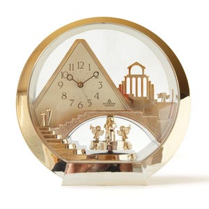 May include: A gold-toned quartz clock with a clear, round face. The clock features a pyramid-shaped face with Roman numerals, a bridge, and angel figurines. The clock is set on a silver base.