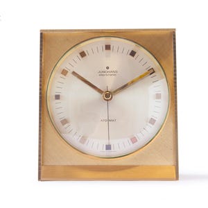 May include: A gold-toned, square-shaped Junghans electronic Ato Mat clock. The clock face is cream-colored with gold hands and small colored squares marking the hours. The clock has the text "Junghans electronic" and "Ato Mat".