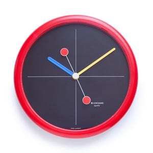 Könnte beinhalten: Eine runde Wanduhr mit rotem Rahmen und schwarzem Zifferblatt. Die Uhr hat einen gelben Stundenzeiger, einen blauen Minutenzeiger und einen weißen Sekundenzeiger. Zwei rote Kreise und weiße Linien ergänzen das Design. Der Text "JUNHANS quartz" ist zu sehen.