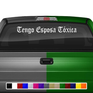 May include: A white truck with a green tailgate. The rear window has a white decal that reads "Tengo Esposa Tóxica" in a gothic font.
