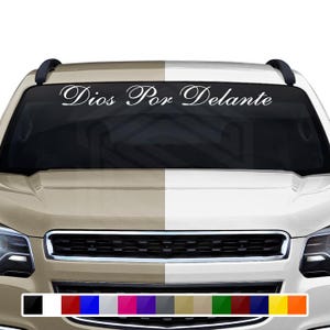 Puede incluir: Una calcomanía de vinilo blanco con el texto "Dios Por Delante" en una elegante escritura, diseñada para ser aplicada en el parabrisas de un vehículo. La imagen muestra la calcomanía aplicada en la parte superior del parabrisas de un coche.
