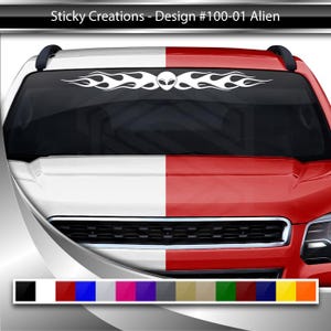 May include: A white alien face with flames on either side, on a red and white truck windshield. The truck is a newer model with a black grill and headlights.