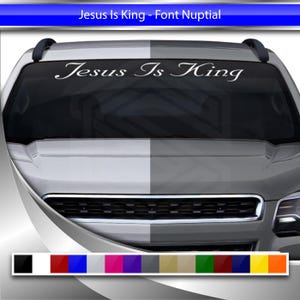 May include: A white car with a windshield decal that reads "Jesus Is King" in a cursive font. The decal is on the driver's side of the windshield. The car is a light gray color and has a black grille.