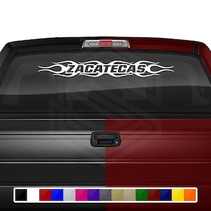 May include: White decal with the word "ZACATECAS" in a stylized font, framed by flame-like designs. The decal is affixed to the rear window of a vehicle, with a black and red color scheme.