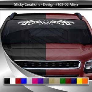 May include: A white alien head graphic with tribal-style flames on either side. The graphic is on a black and red car windshield.