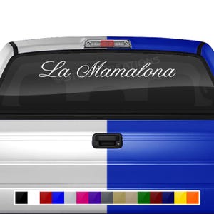 May include: A white truck with a blue tailgate. The rear window has a white vinyl decal that reads "La Mamalona".